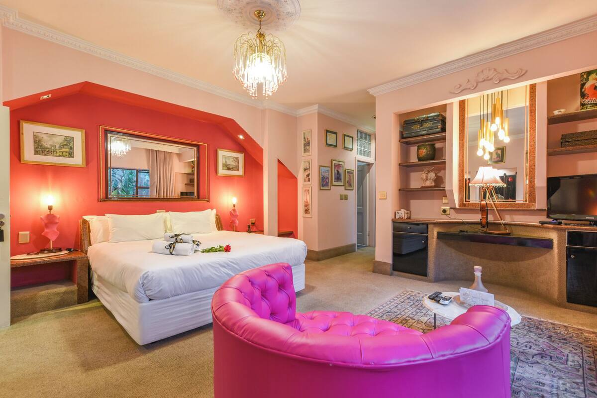 Una habitación de hotel con una cama, un reposapiés rosa, un escritorio con televisor y una araña.