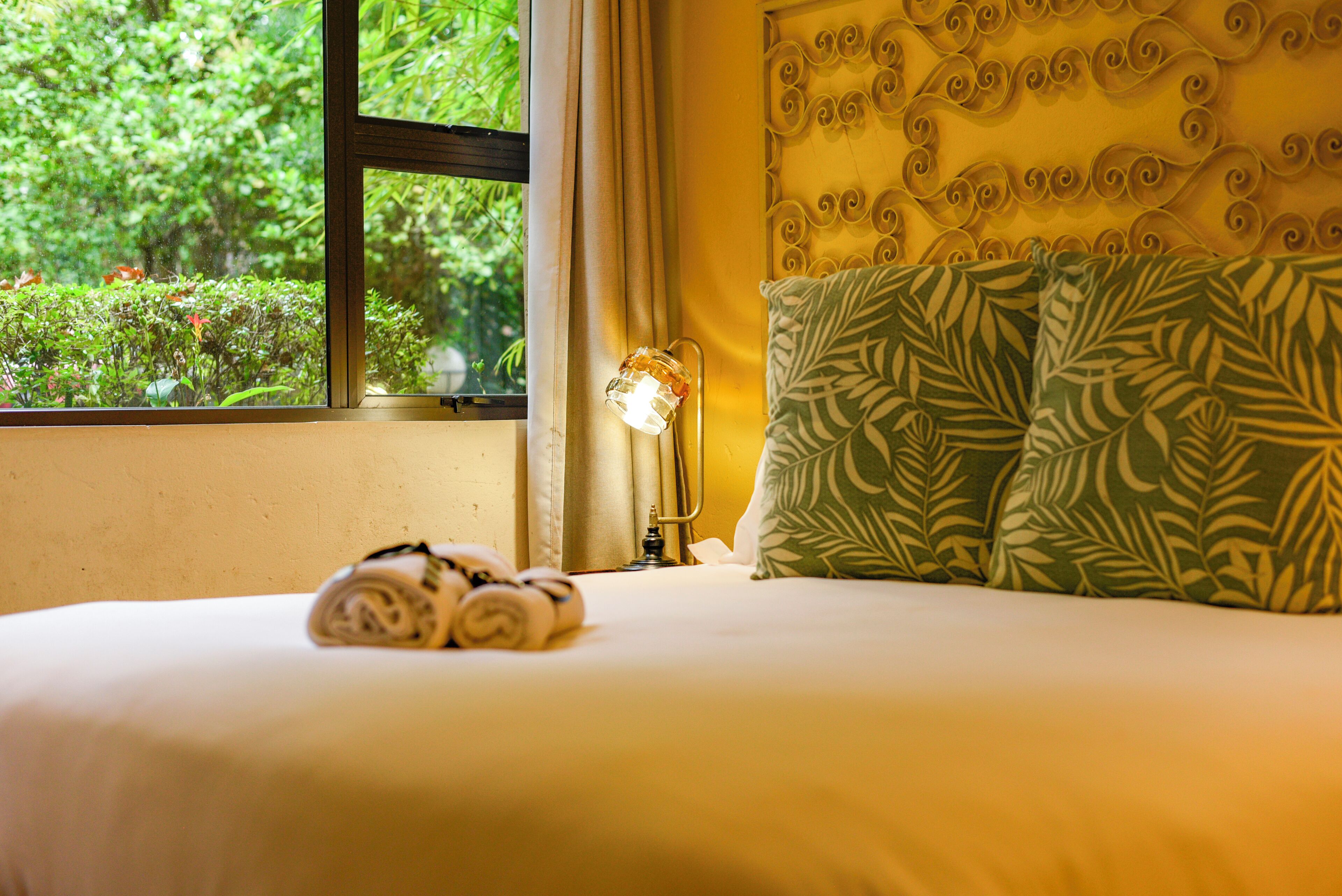 A hotel room with a bed, a window with curtains, a lamp, and a view of greenery outside.