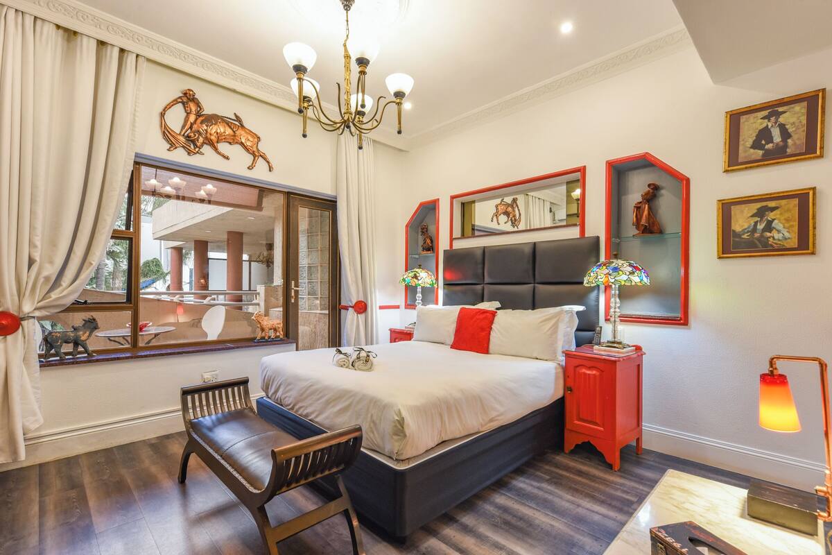 Un dormitorio con una cama grande, una mesita de noche, una silla, una araña y una ventana con cortinas.