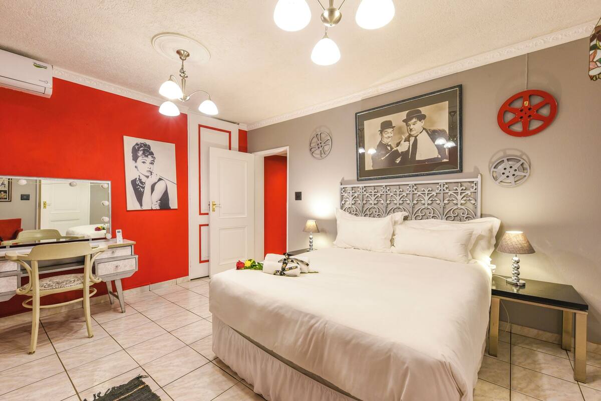 Una habitación de hotel con cama, mesita de noche, lámpara, cómoda, espejo y una pared decorada en rojo.