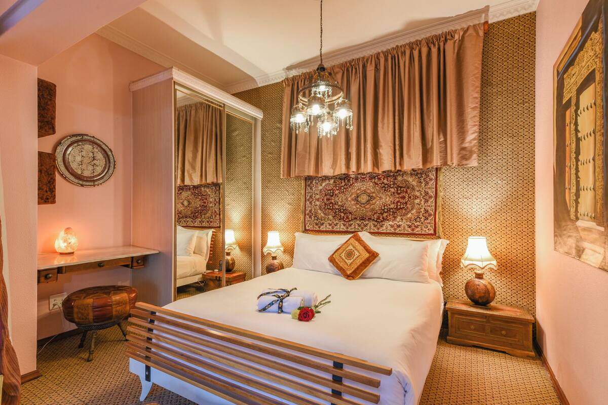 Habitación de hotel con cama, lámparas de noche, mesita de noche, espejo, banco de madera y decoración en la pared.