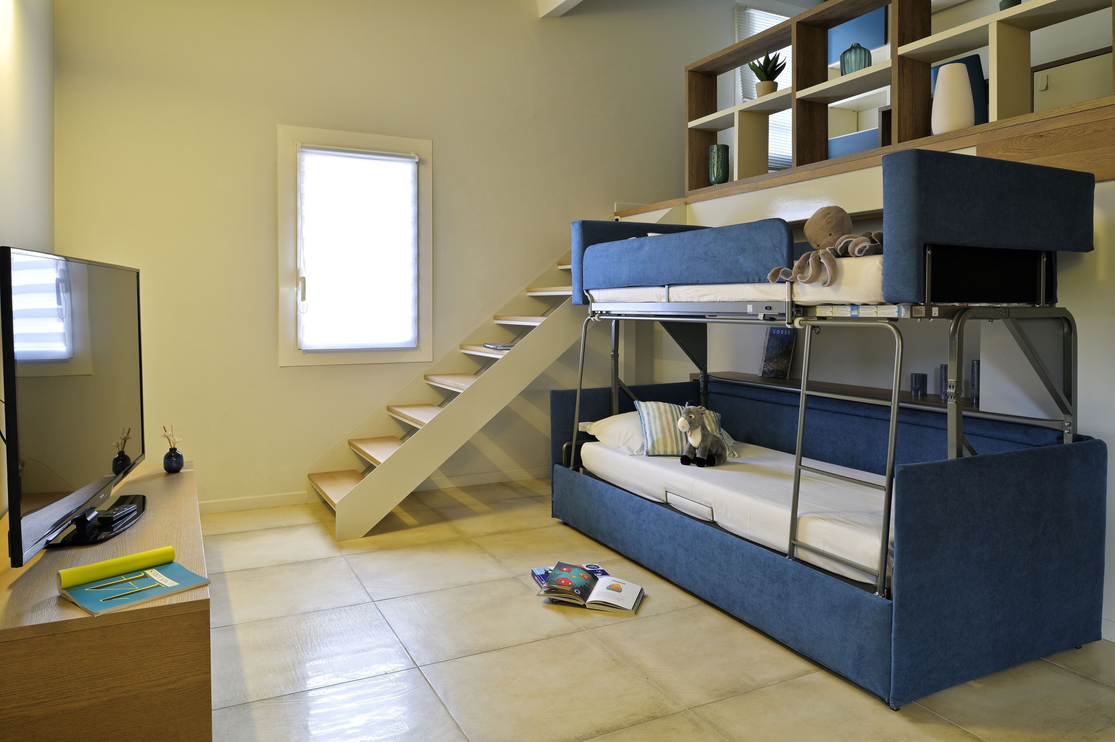 Ein kleines Zimmer mit Etagenbett, Fernseher, Schreibtisch und Treppe.