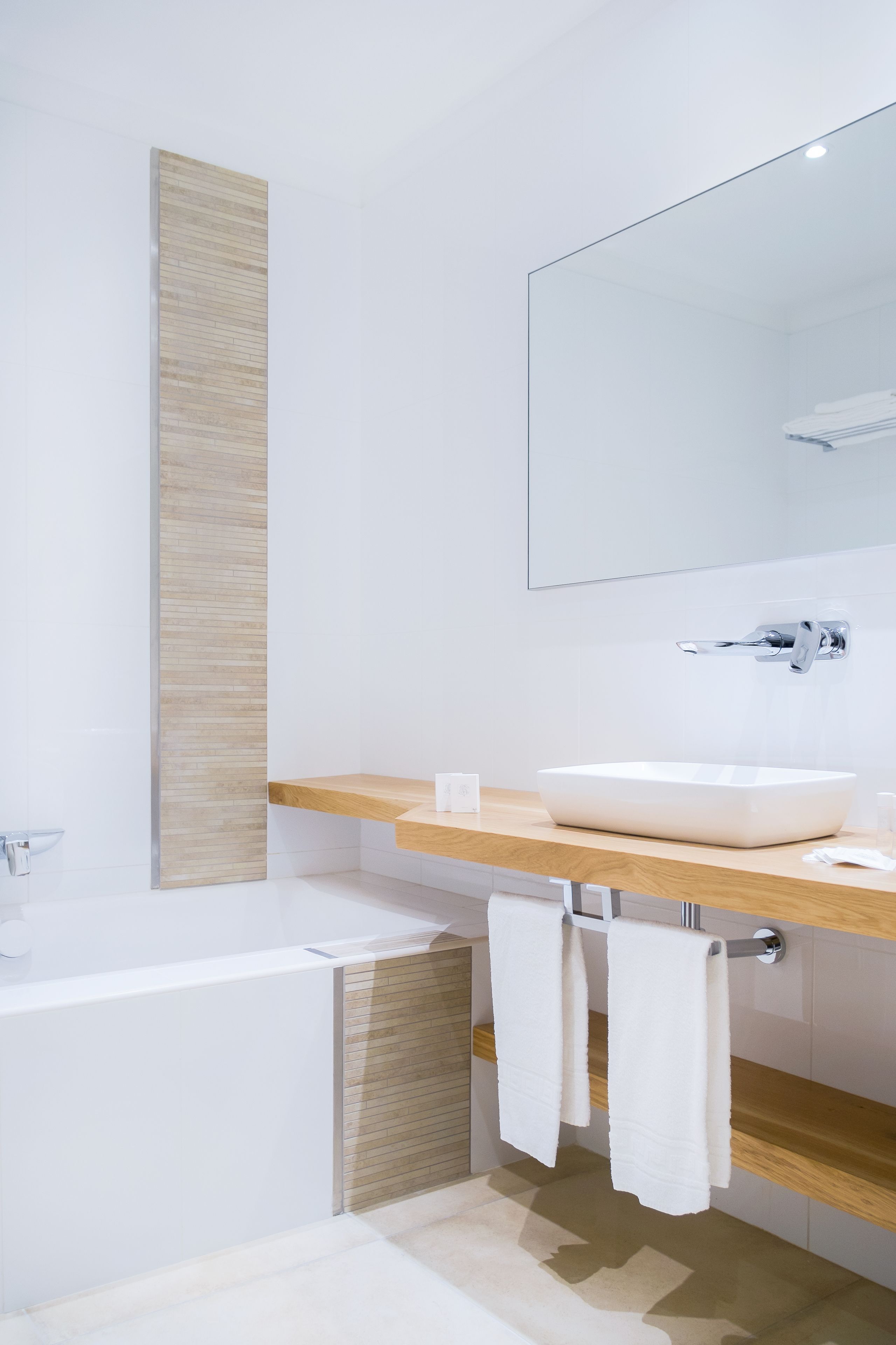 Une salle de bain moderne avec une baignoire blanche, un meuble-lavabo en bois, un grand miroir et deux serviettes blanches suspendues au porte-serviettes.