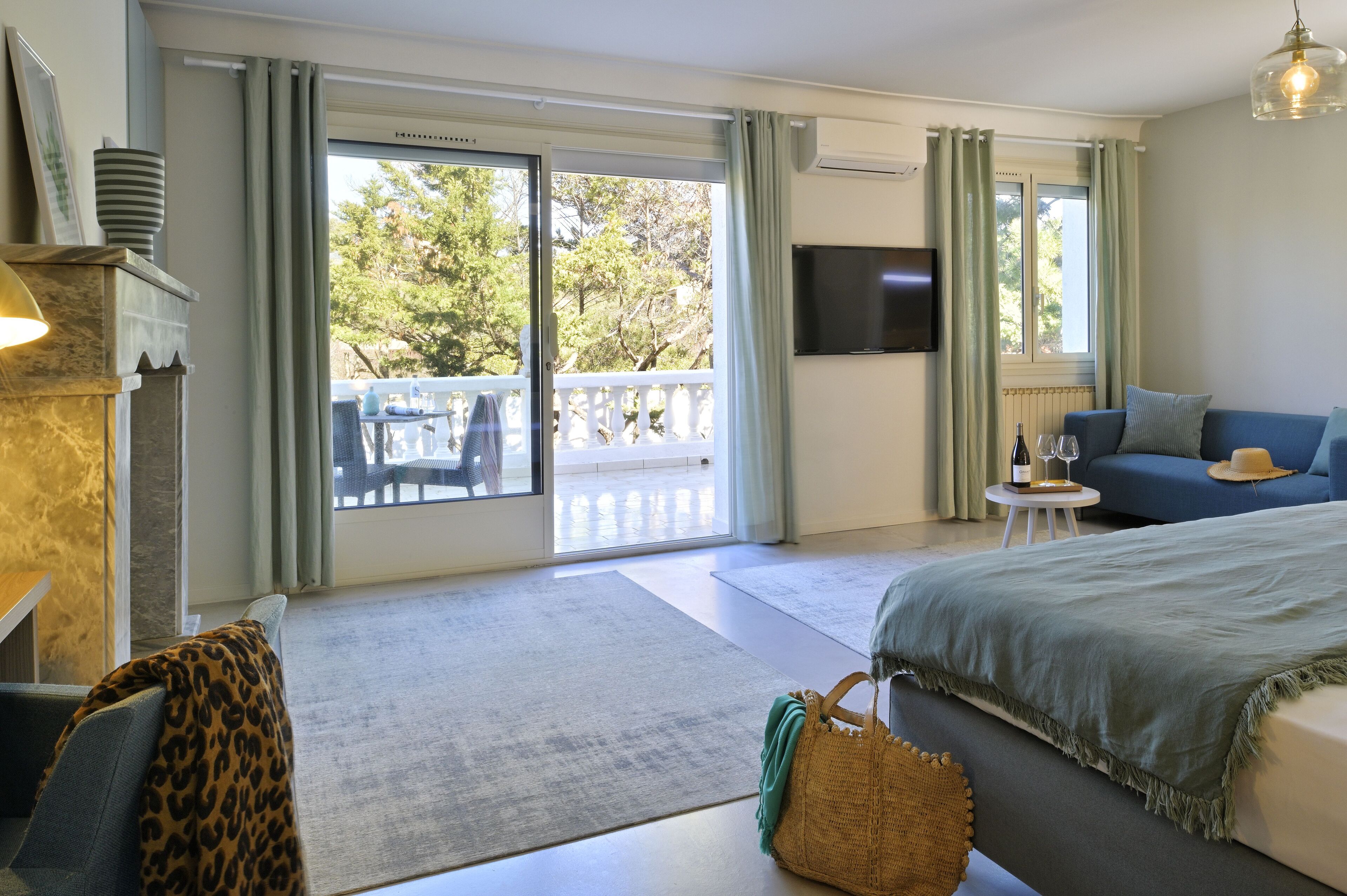 A hotel room with a large bed, a sofa, a TV, a balcony with a view of trees, and a fireplace.