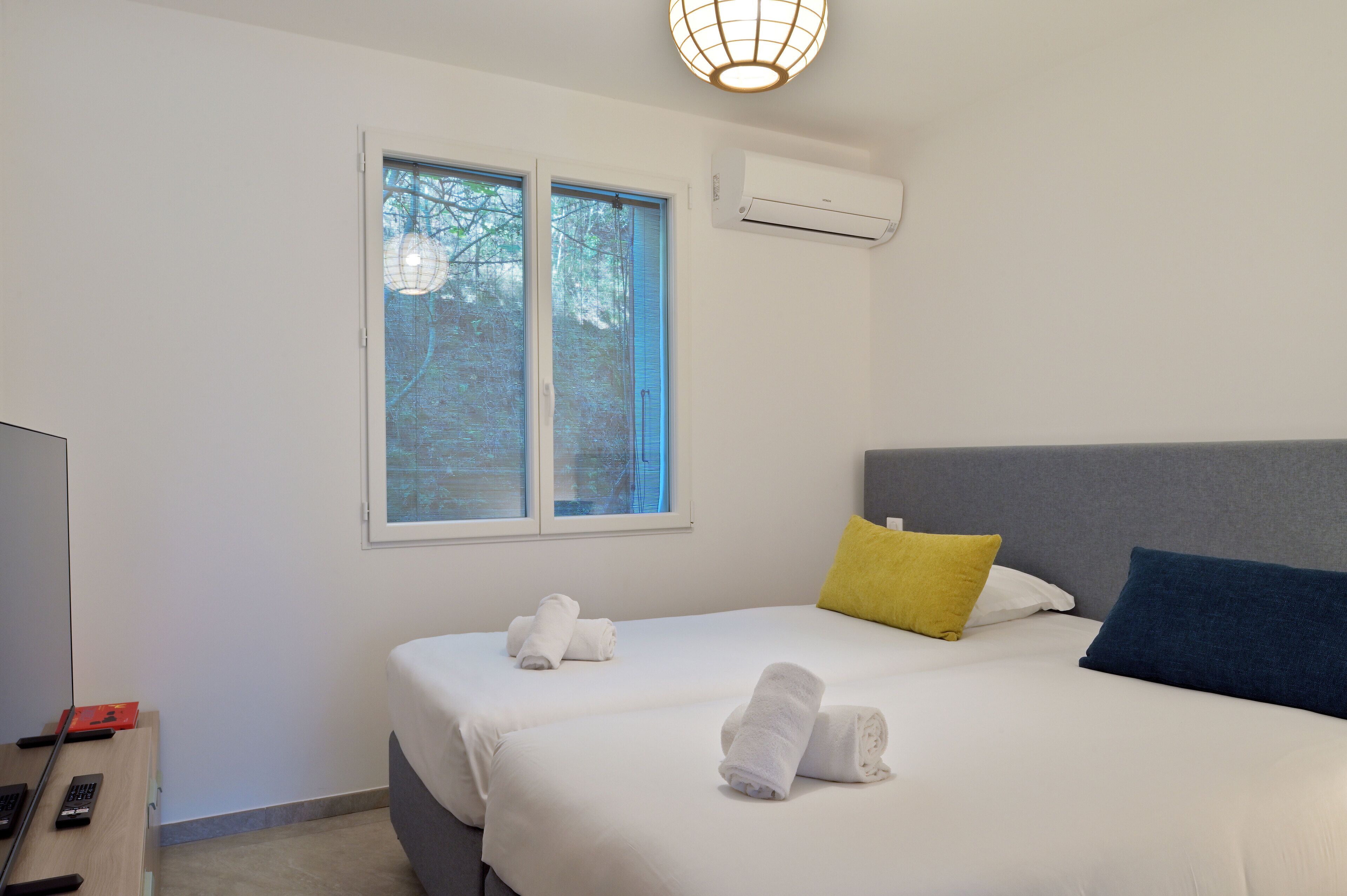 A hotel room with a bed, two pillows, a bedside table with a remote control, and an air conditioning unit.