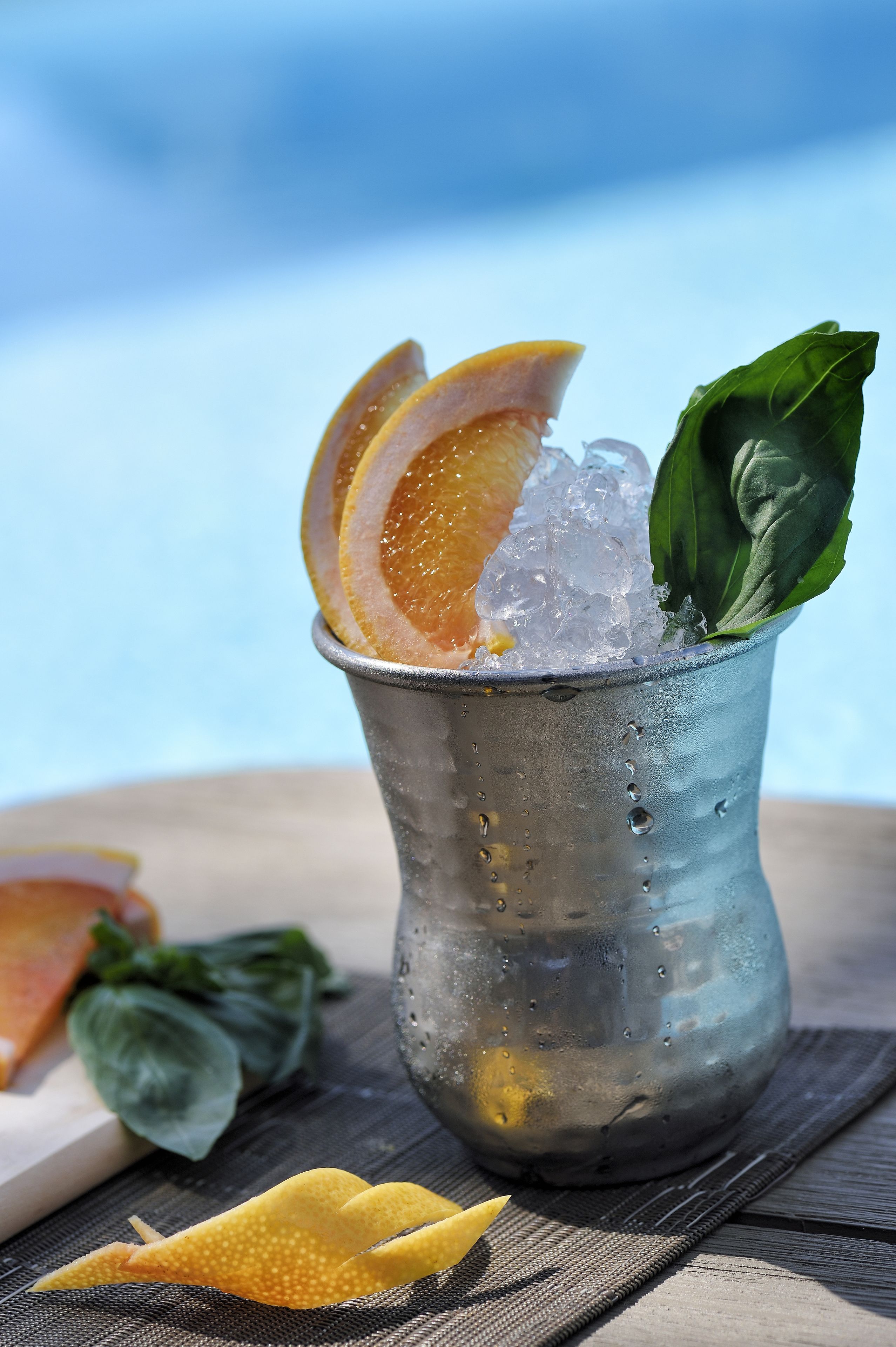 Ein Metallbecher gefüllt mit Eiswürfeln, Orangenscheiben und einem grünen Blatt, auf einer Holzunterlage vor unscharfem, blauem Hintergrund.