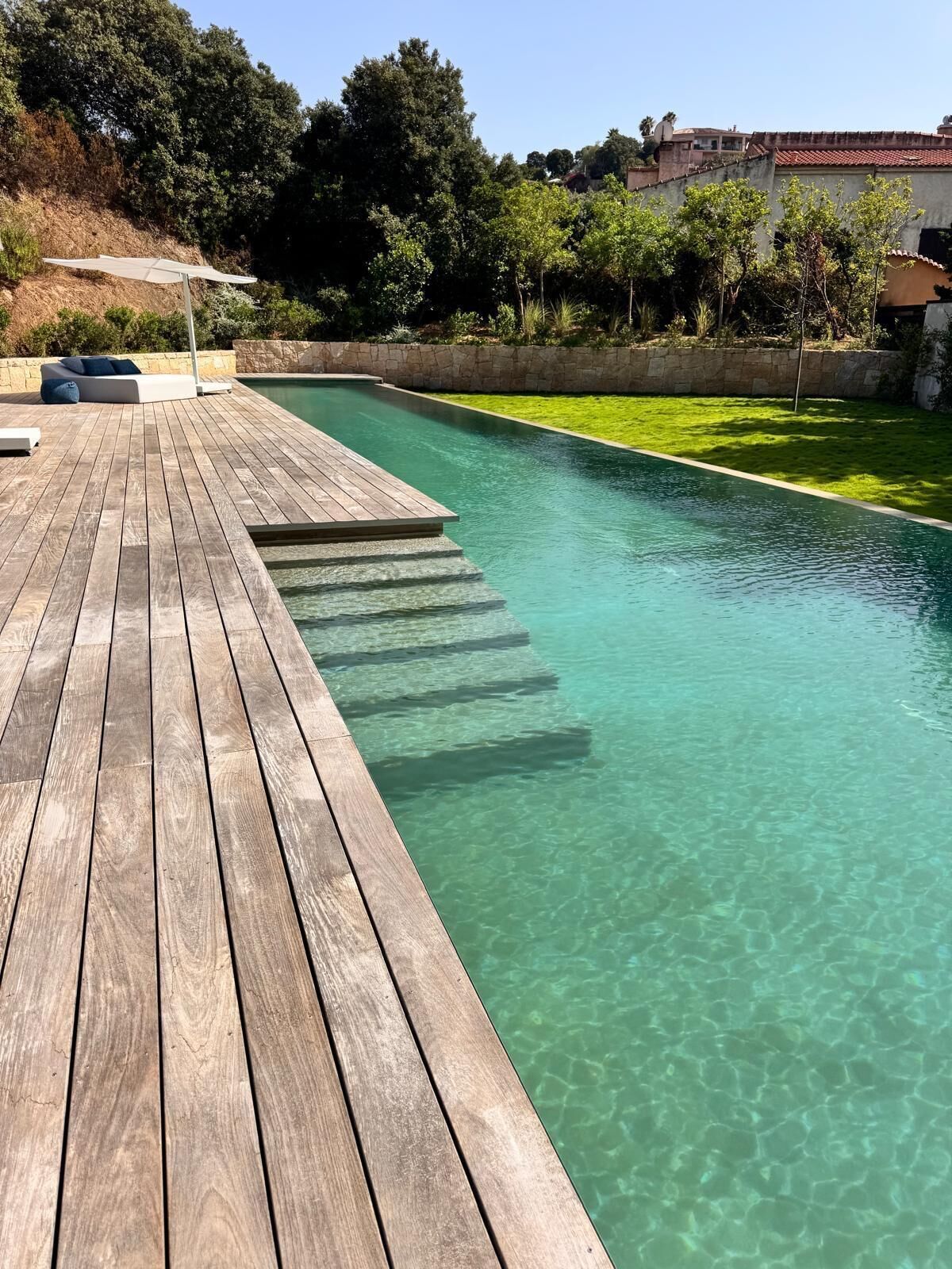 A wooden deck leading to a clear swimming pool with a grassy area and trees in the background.