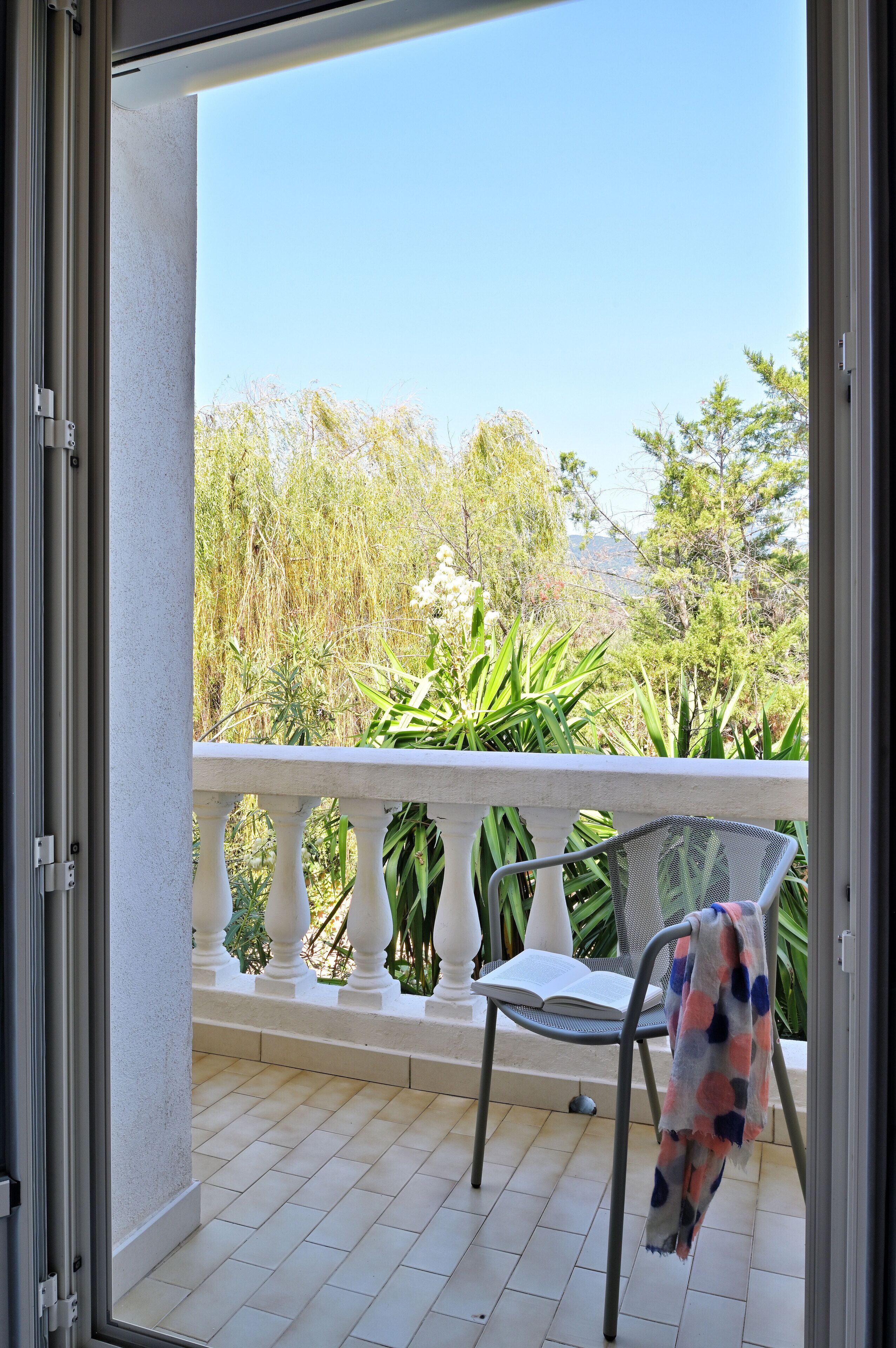 Een balkon met een witte balustrade, een metalen stoel met een deken, en uitzicht op groen en bomen.
