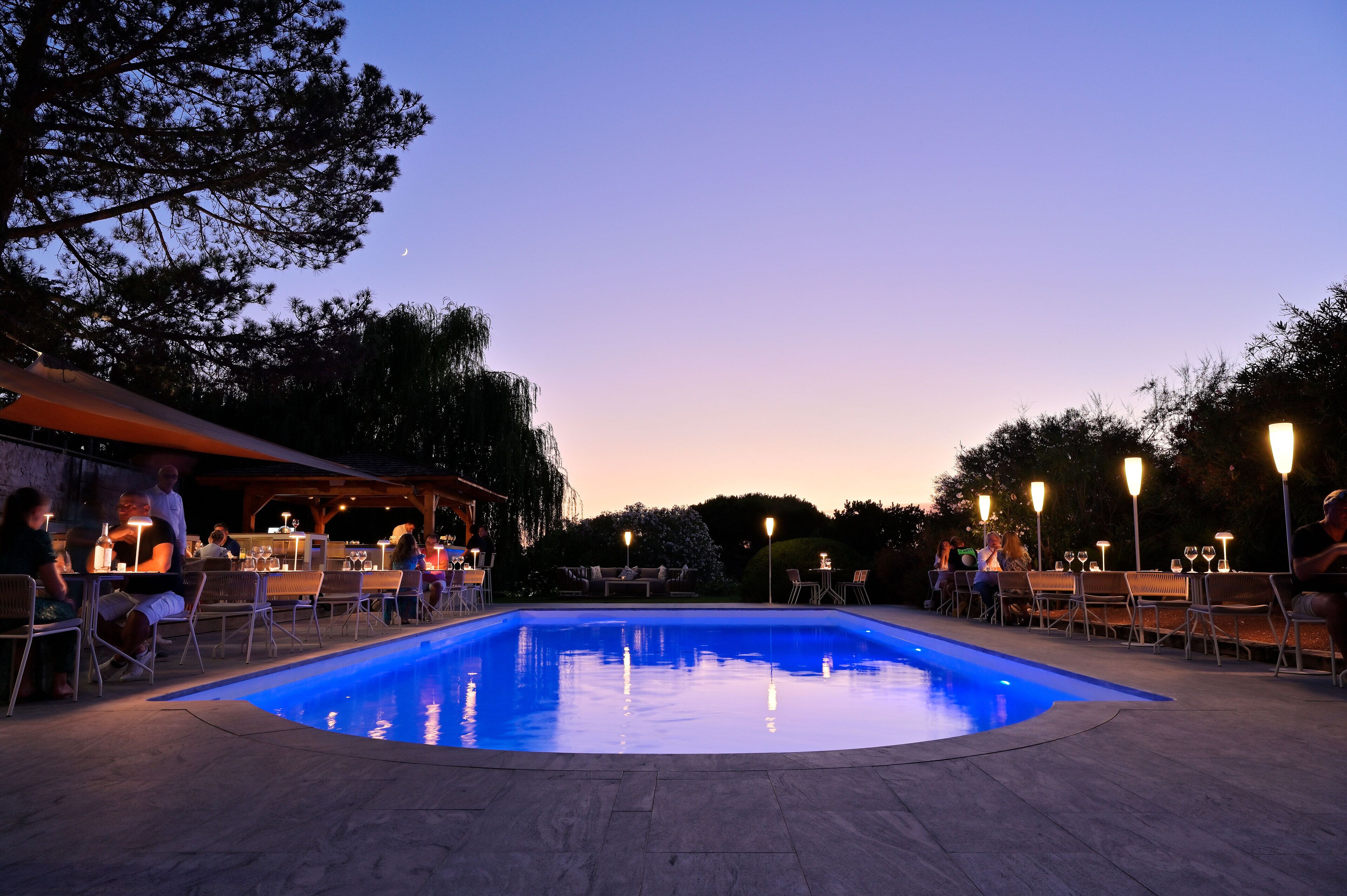 An outdoor pool area with seating and tables, surrounded by trees and a bar area.