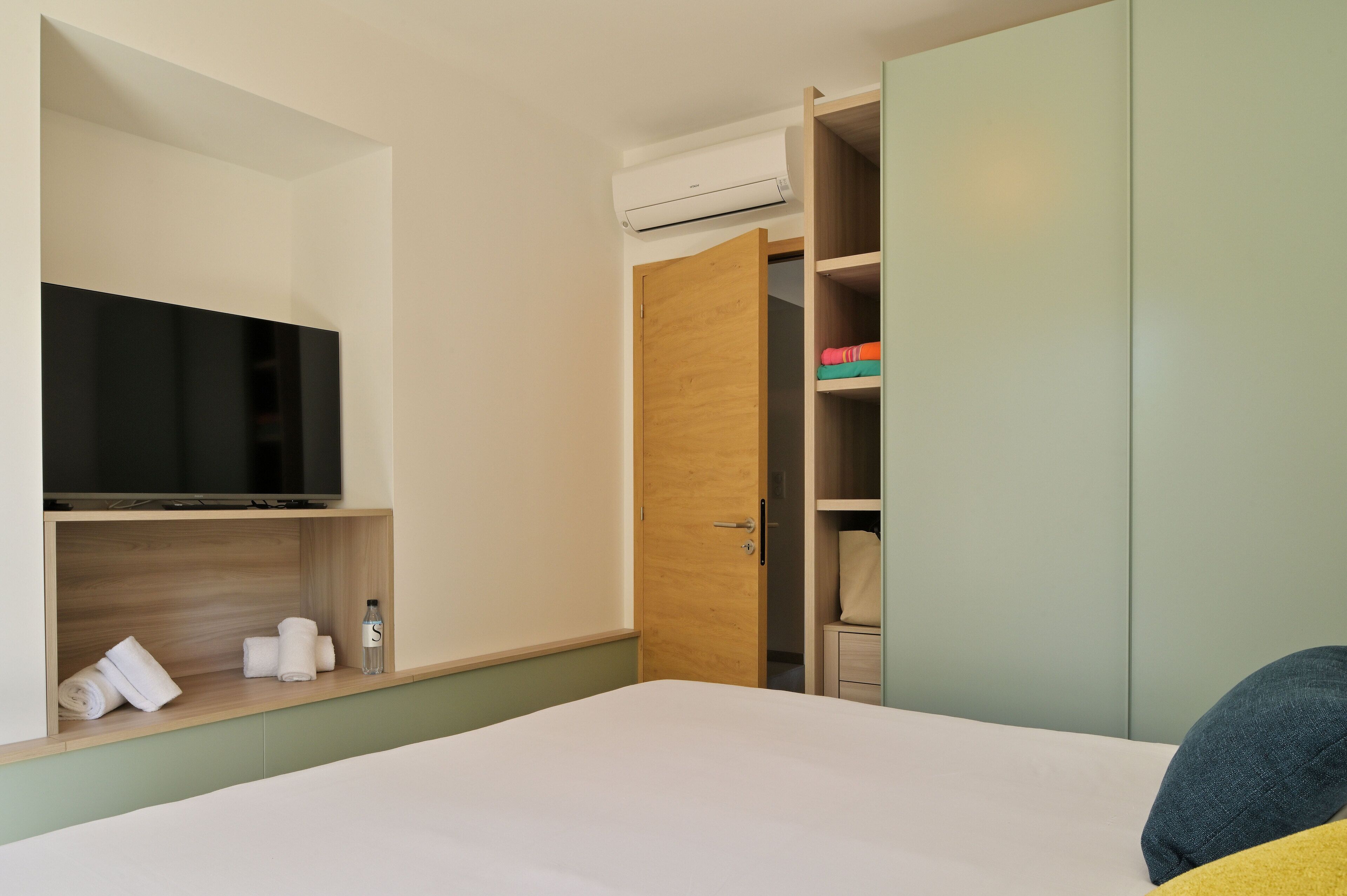 A hotel room with a bed, a TV on a shelf, a wardrobe, and a closet with clothes.