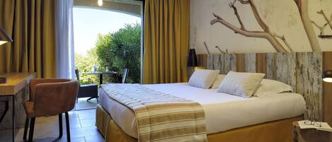 A hotel room with a large bed, a desk, and a chair. There is a view of greenery outside the window.
