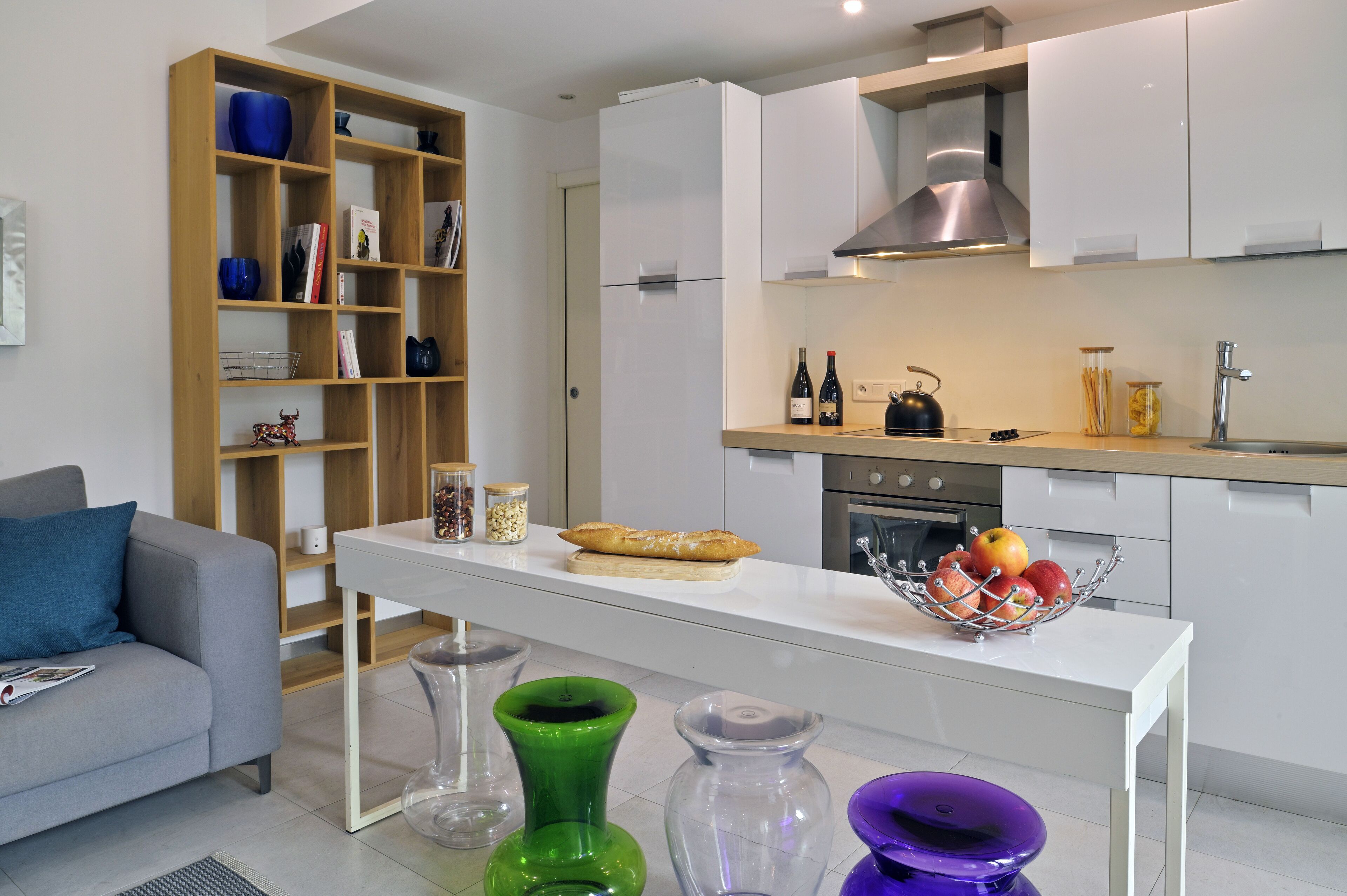 A modern kitchen with white cabinets, a wooden bookshelf, a dining table with a bread basket, and a fruit bowl.