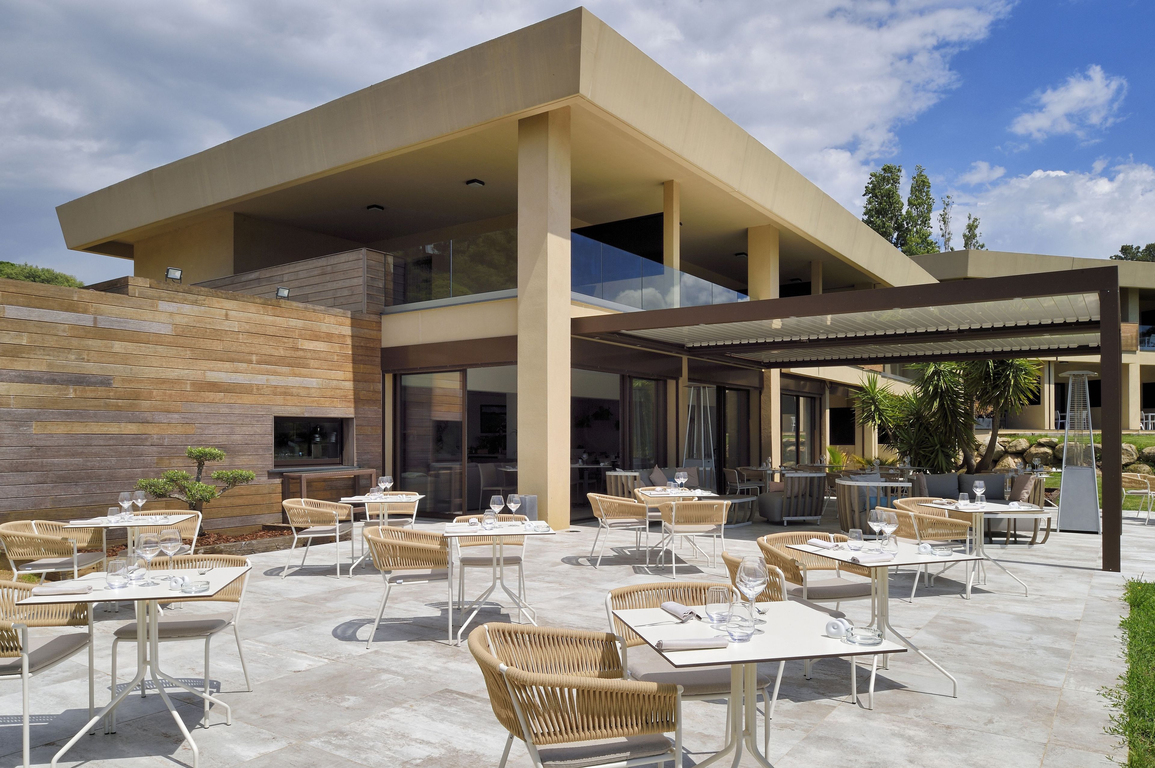 An outdoor dining area with tables and chairs, a covered seating area, and a modern building with large windows and a wooden wall.