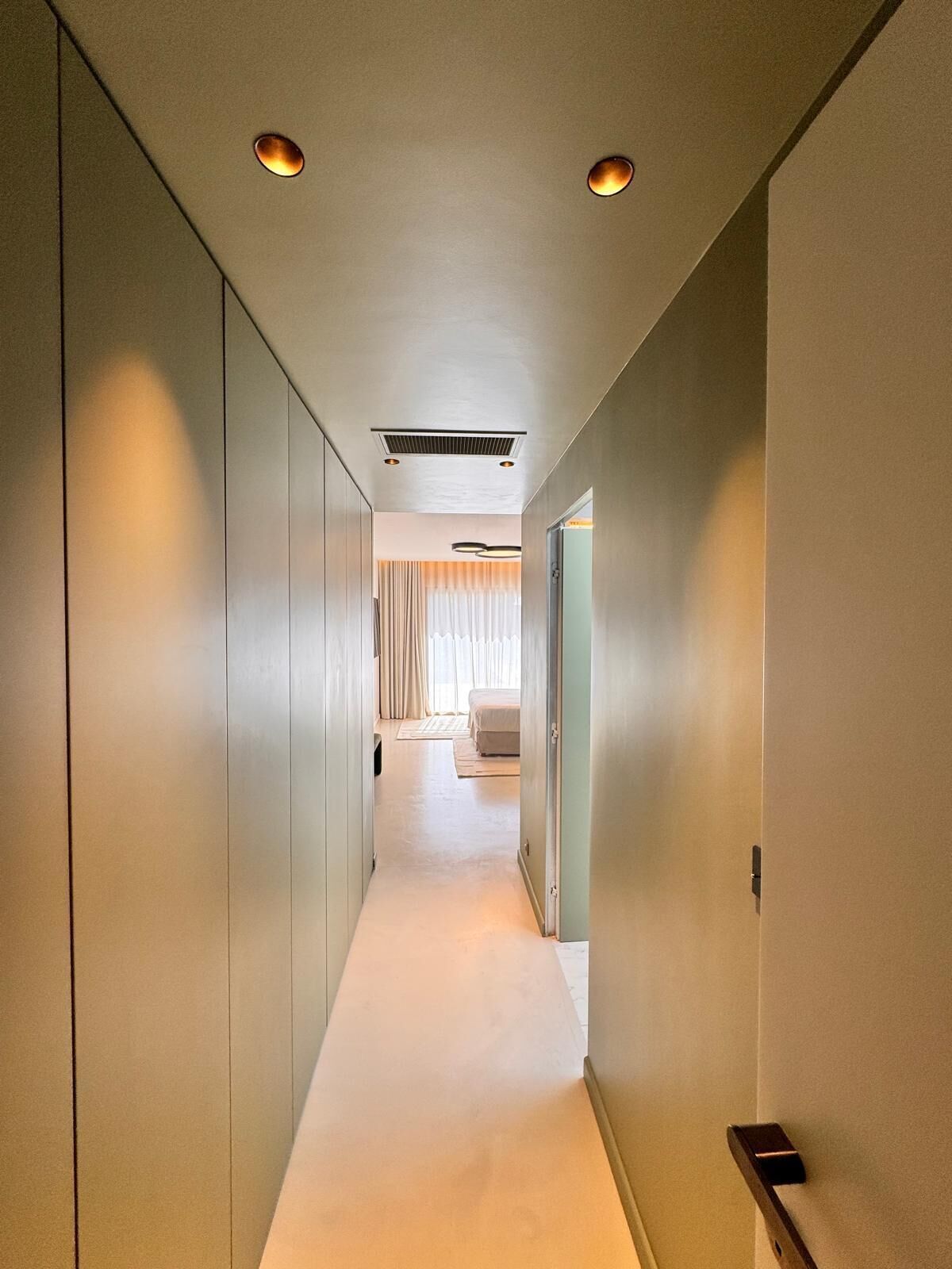 A hallway with a light-colored carpet, a ceiling with recessed lighting, and a door leading to a room with a bed and curtains.