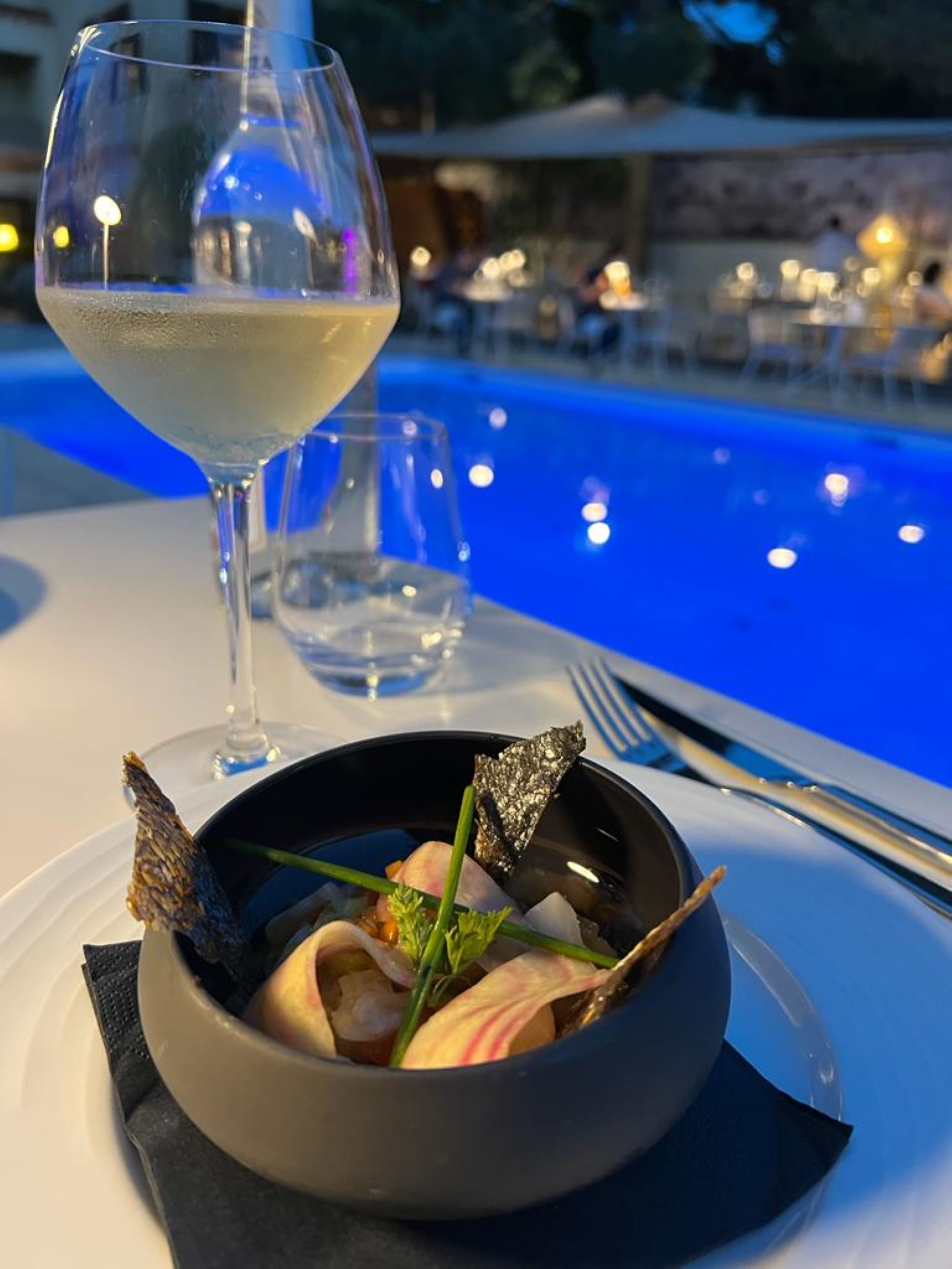 A dining table with a bowl of seafood, a wine glass, and a poolside view.