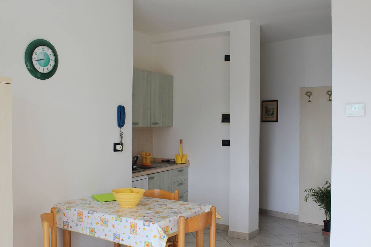 Zona pranzo con tavolo apparecchiato per due, cucina sullo sfondo e una pianta in vaso a destra.