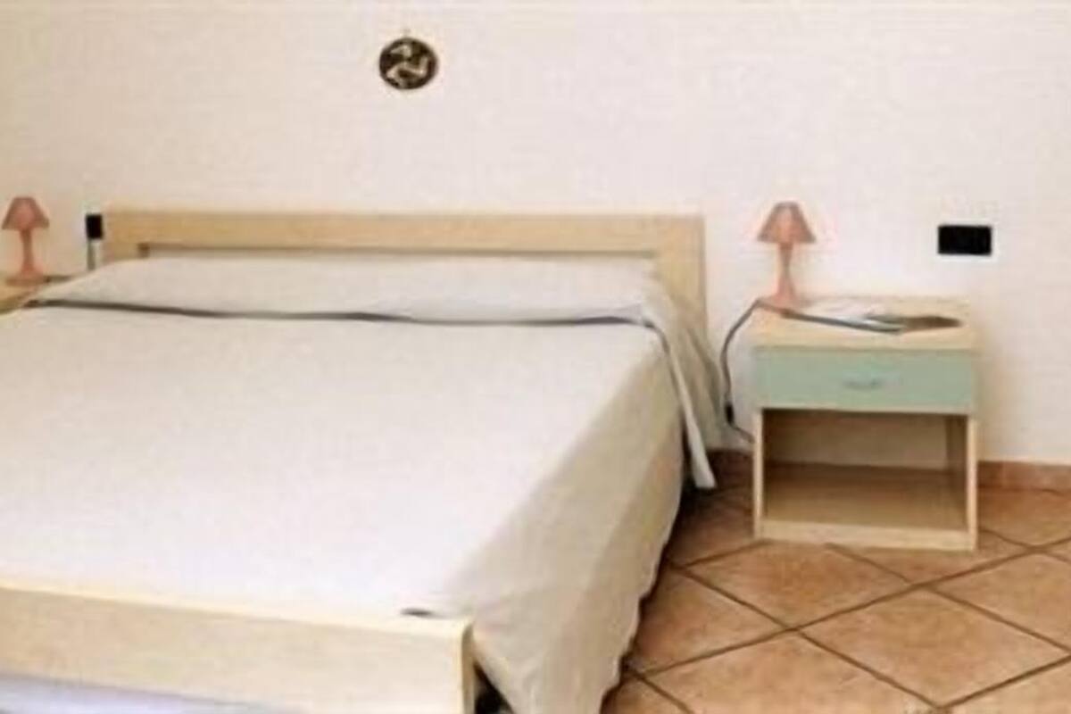 Camera singola con un letto e una testiera in legno, un comodino con una lampada e un piccolo mobiletto contenitore.