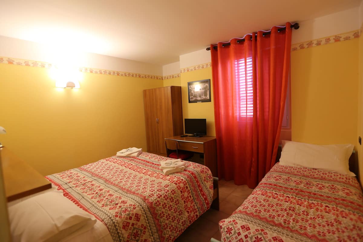 A hotel room with two beds, a desk with a computer, a wardrobe, and a window with red curtains.