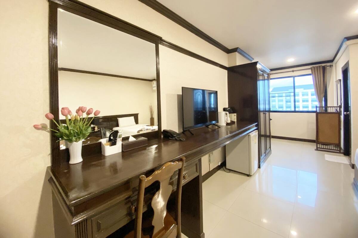 A hotel room with a large mirror, a desk with a telephone, a television, and a window with a view of buildings.
