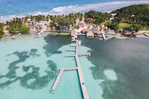 Vista aérea de un resort en una isla tropical, con un muelle que se adentra en aguas turquesas, rodeado de palmeras y casas de playa.