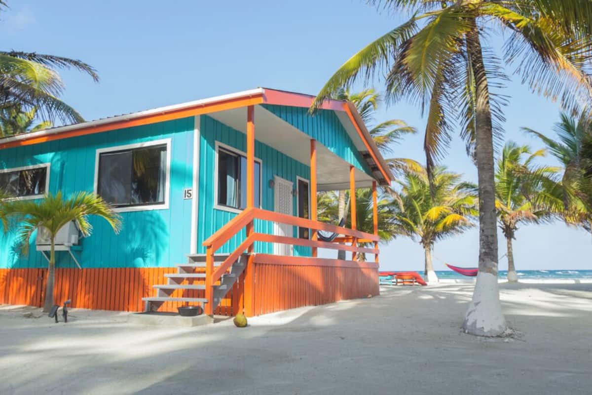 Una casa de playa turquesa y naranja con porche, palmeras y vista al océano.