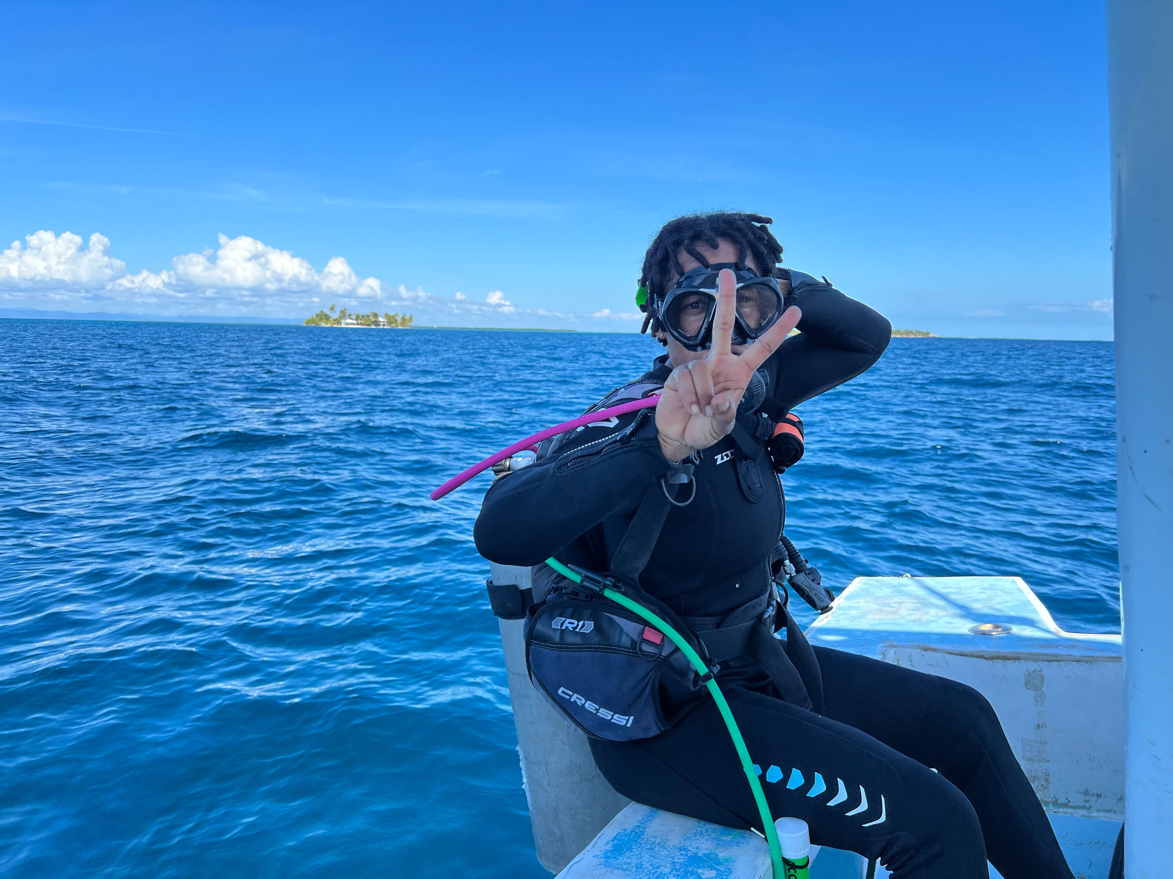 A diver in a black wetsuit with a scuba mask and fins, sitting on a boat with a clear view of the ocean and a distant island.