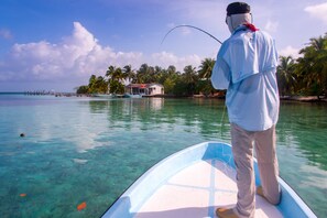 Una persona pescando desde un bote en aguas cristalinas, con una casa y palmeras al fondo.