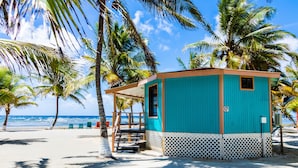 Un petit chalet de plage turquoise, doté d’un petit porche et d’une plaque portant le numéro « 17 », entouré de palmiers et donnant sur l’océan.