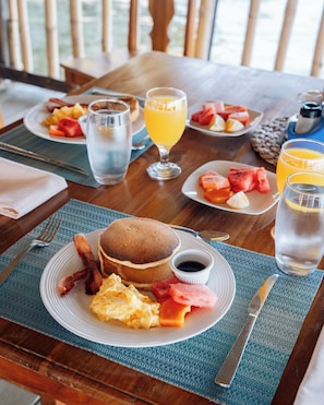 Desayuno con tortitas, tocino, huevos y fruta sobre una mesa de madera.