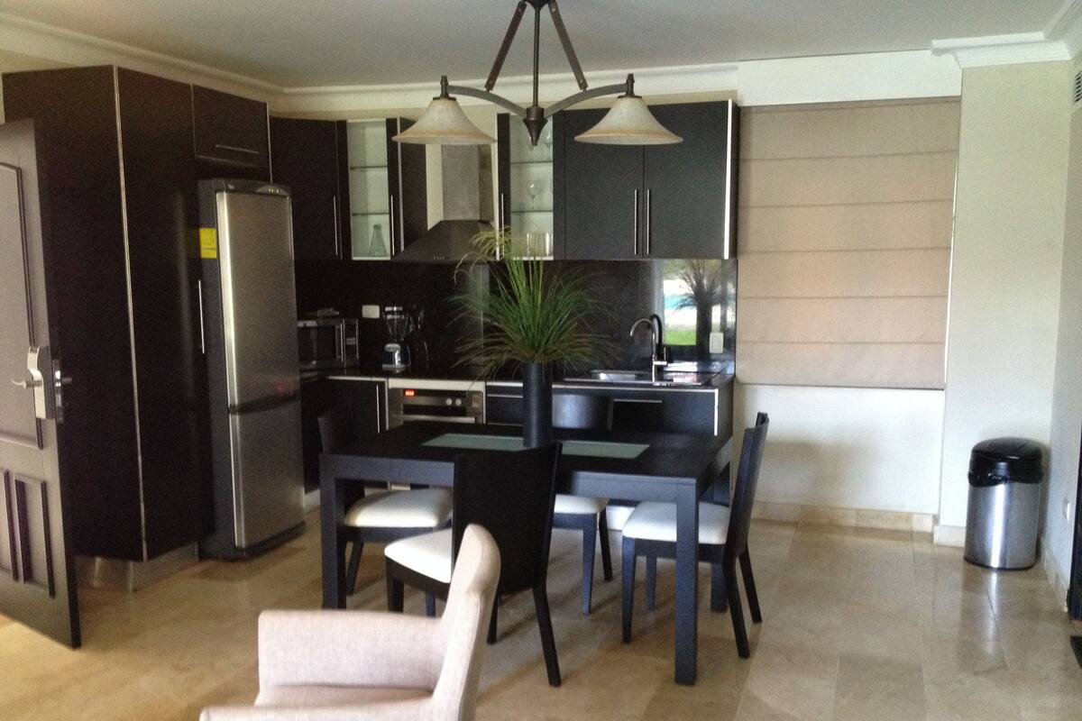 Una cocina moderna con armarios oscuros, un refrigerador de acero inoxidable, una mesa de comedor con sillas y una lámpara colgante.