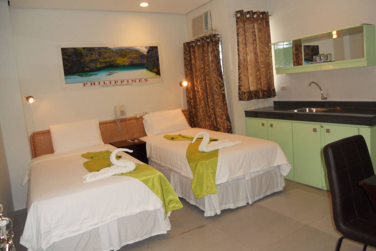 Quarto de hotel com duas camas, uma kitchenette pequena, um banheiro e uma pintura das Filipinas na parede.