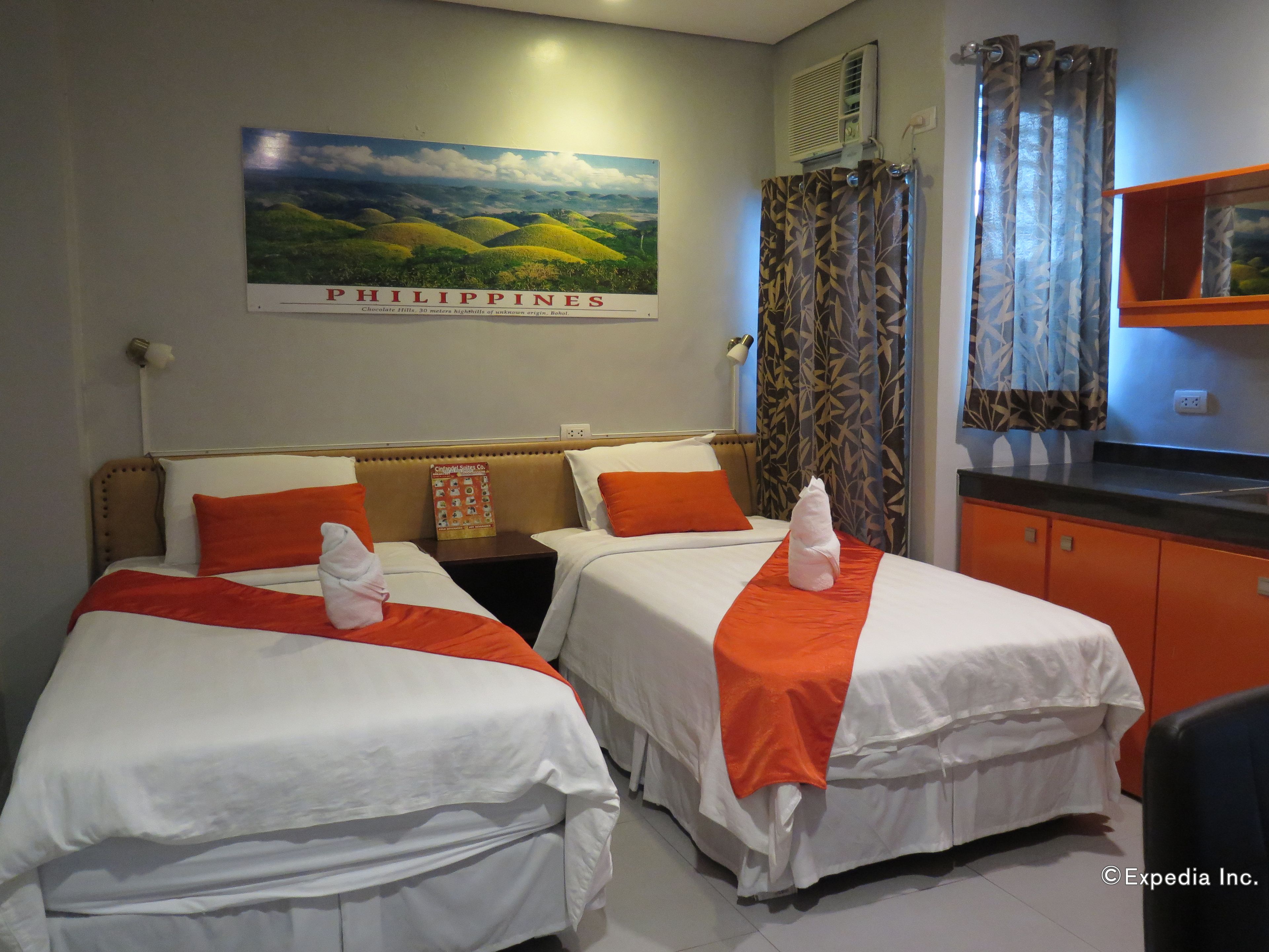 A hotel room with two beds, a nightstand, a window with patterned curtains, and a wall-mounted air conditioner.