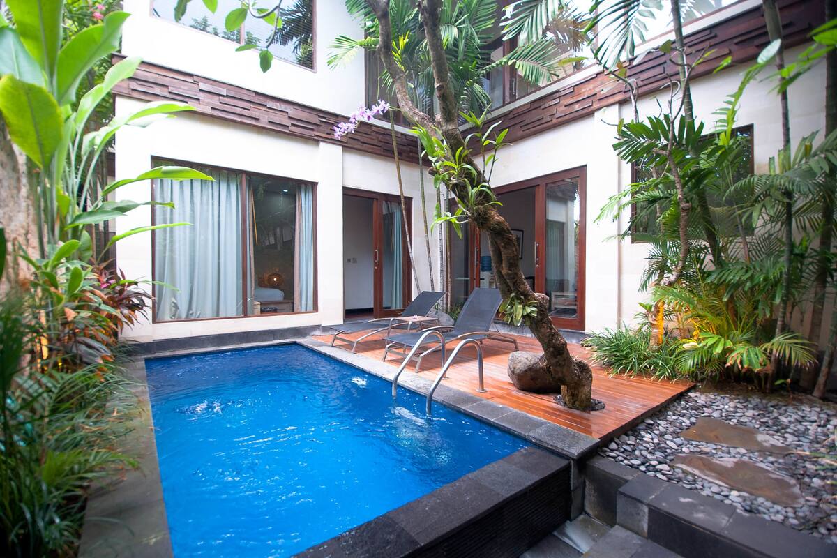 Un área de piscina con terraza de madera, sillas de descanso y plantas tropicales.