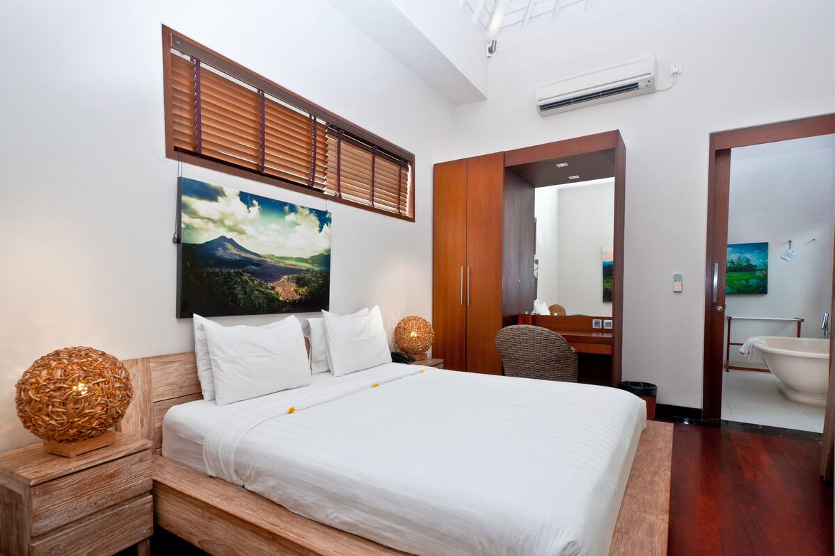 Un dormitorio con una cama grande, cabecera de madera, un cuadro de un paisaje, una mesita de noche de madera, una lámpara de mimbre, una silla y un baño con bañera y lavamanos.