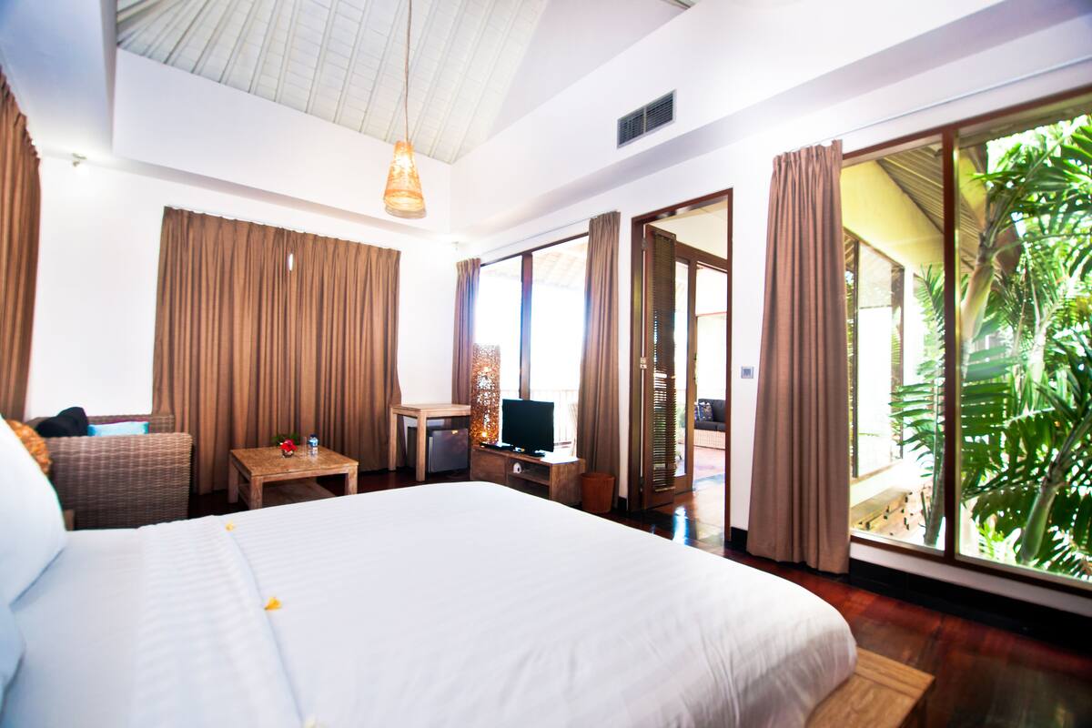 Amplia habitación de hotel con una cama grande, un escritorio, un televisor y vistas a un follaje exuberante a través de las ventanas.