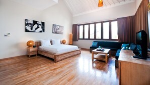 A spacious room with a large bed, a sofa, a coffee table, and a television. There are two paintings on the wall, a window with curtains, and a ceiling with a grid pattern. - The Dipan Resort Petitenget (Keroboken Kelod)