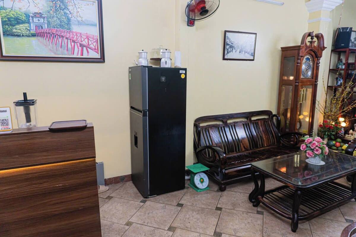 Una habitación con un armario de madera, un refrigerador negro, un sofá de madera, una mesa de centro de vidrio y un ventilador de pared.