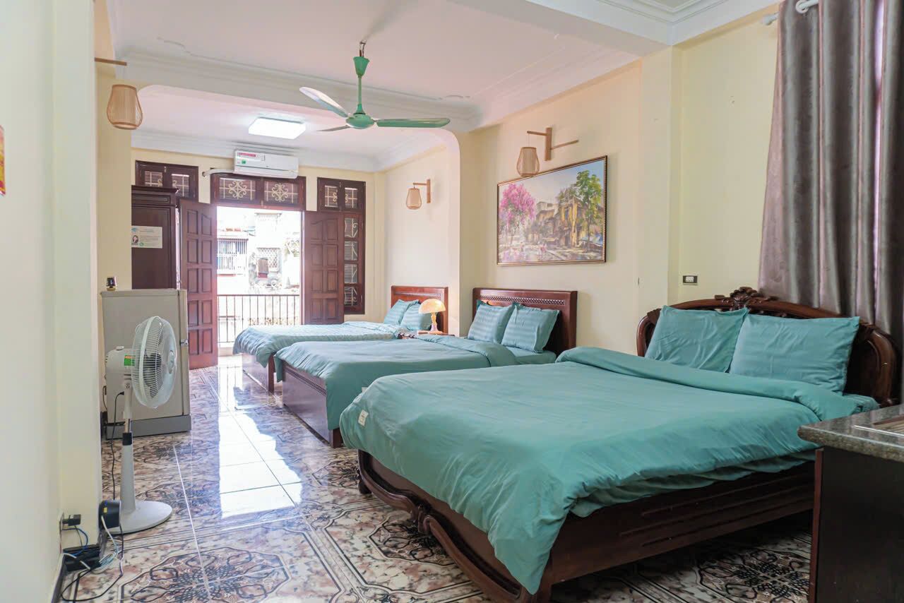 Quarto de hotel com duas camas, um ventilador de teto, um ventilador no chão, um quadro na parede e piso de azulejos.