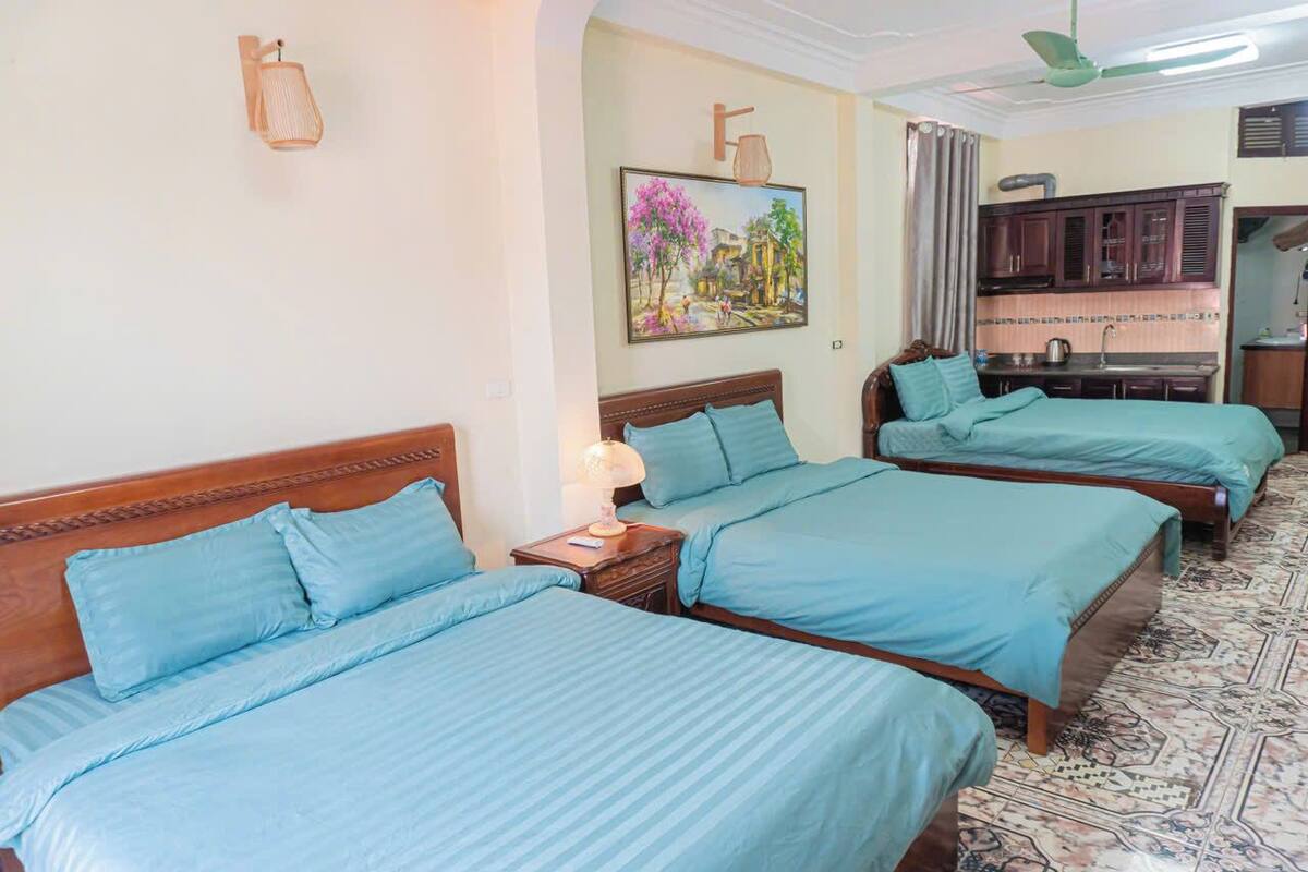 Una habitación de hotel con tres camas, un cuadro en la pared, un ventilador de techo y una zona de cocina.