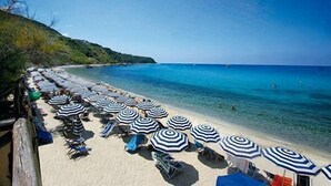 A beach with rows of striped umbrellas and lounge chairs, a clear blue sea, and a hilly landscape in the background. - Baia Tropea Resort (Parghelia)