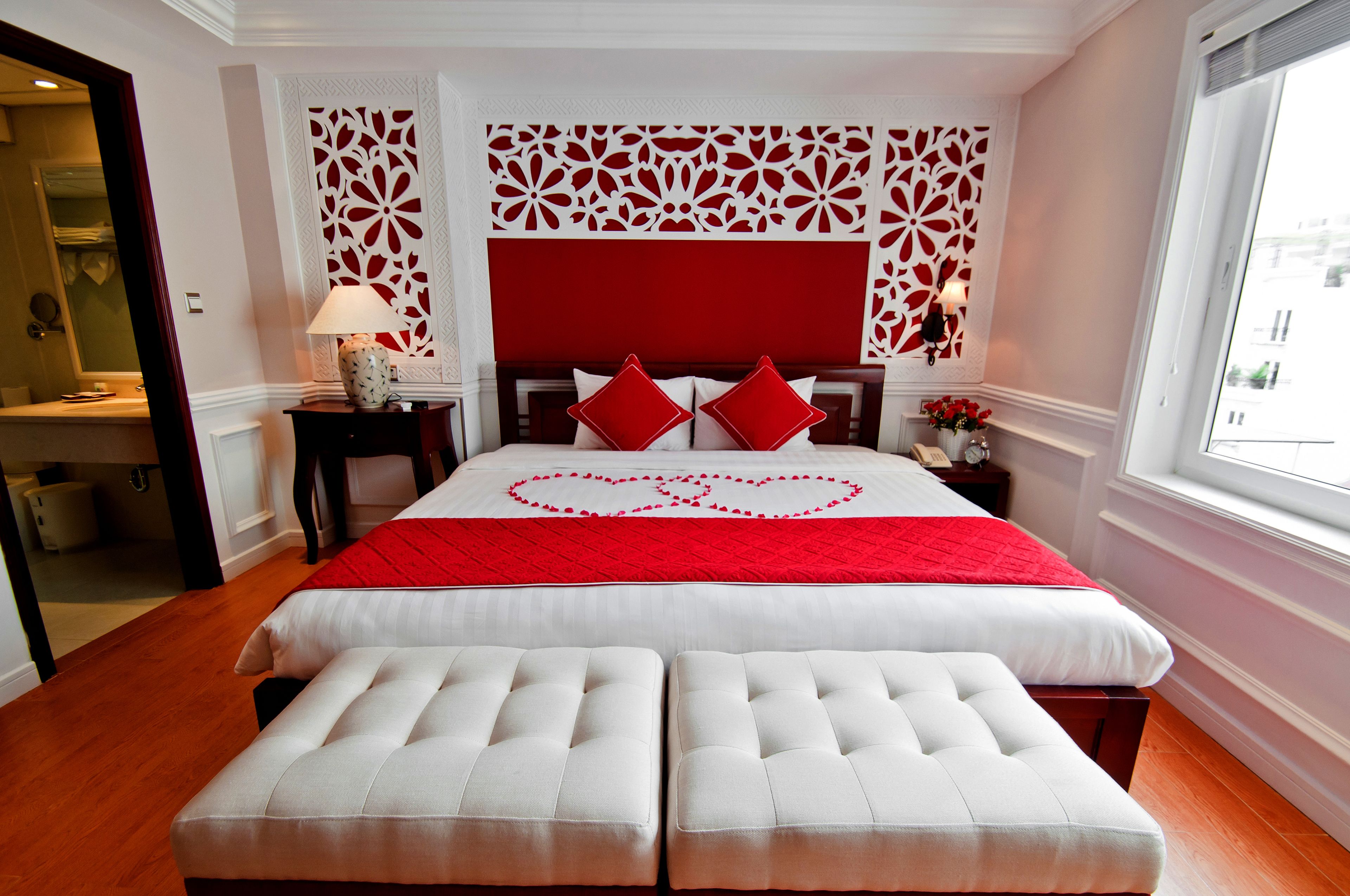 Une chambre d’hôtel avec un grand lit, deux poufs blancs, une décoration en forme de cœur rouge sur le lit, une tête de lit rouge et un panneau décoratif mural.