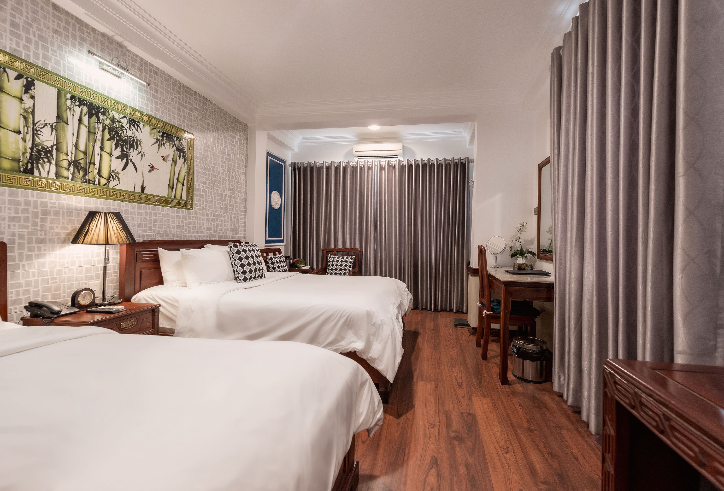 A hotel room with two beds, a desk, a chair, a lamp, a clock, and a painting on the wall.