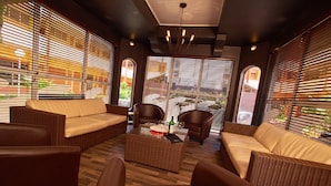 Un espacio lounge moderno con mobiliario de mimbre, una mesa de centro y ventanas amplias con persianas.