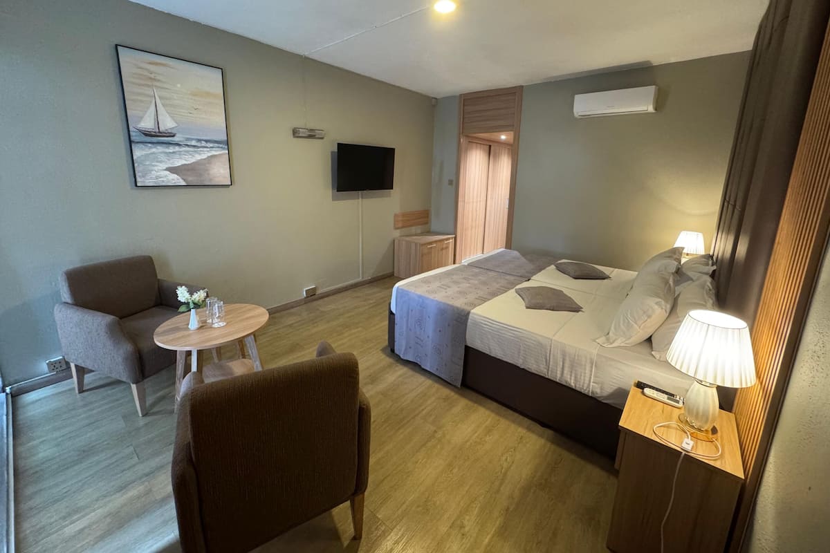 A hotel room with a bed, bedside table with a lamp, a TV, a painting, a chair, and a small table with a vase and glasses.