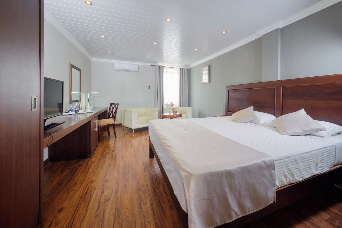 Habitación de hotel con piso de madera, una cama con ropa blanca, un escritorio con una computadora, una silla y una mesita con un florero.