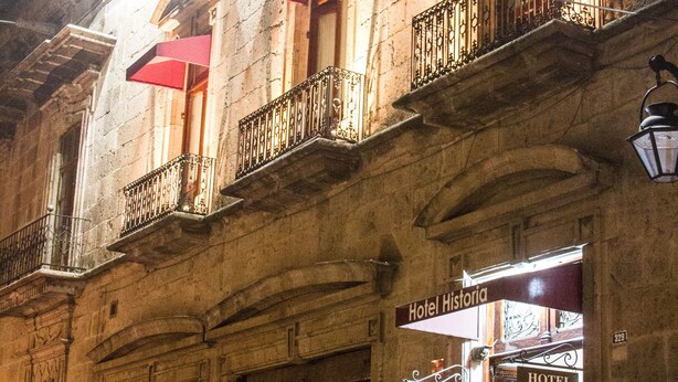 A historic building with a sign for 'Hotel Historia' and closed shutters on the ground floor. - Hotel Historia (Morelia)