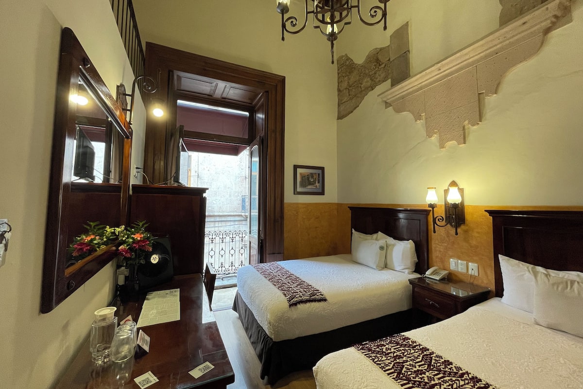Habitación de hotel con dos camas, un aparador de madera, un escritorio con un ventilador, un espejo y un candelabro.