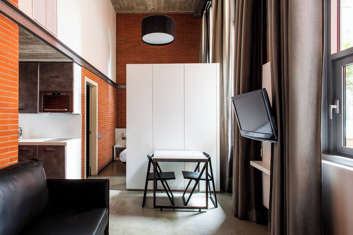 A modern interior with a brick wall, white cabinets, a black leather couch, a small table with chairs, and a TV mounted on the wall.