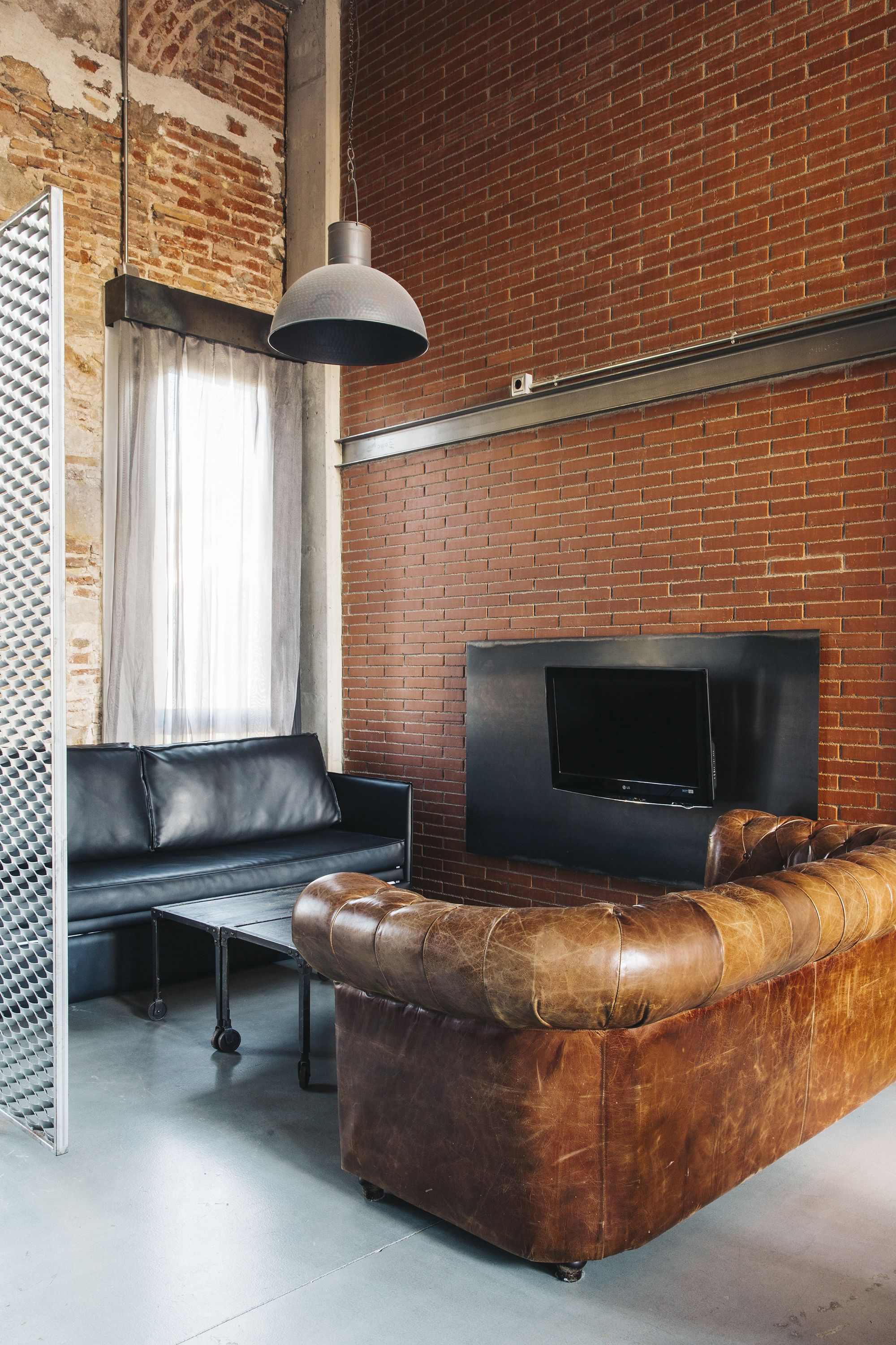A modern living room with a brick wall, a black leather sofa, a black coffee table, a TV mounted on the wall, and a hanging pendant light.