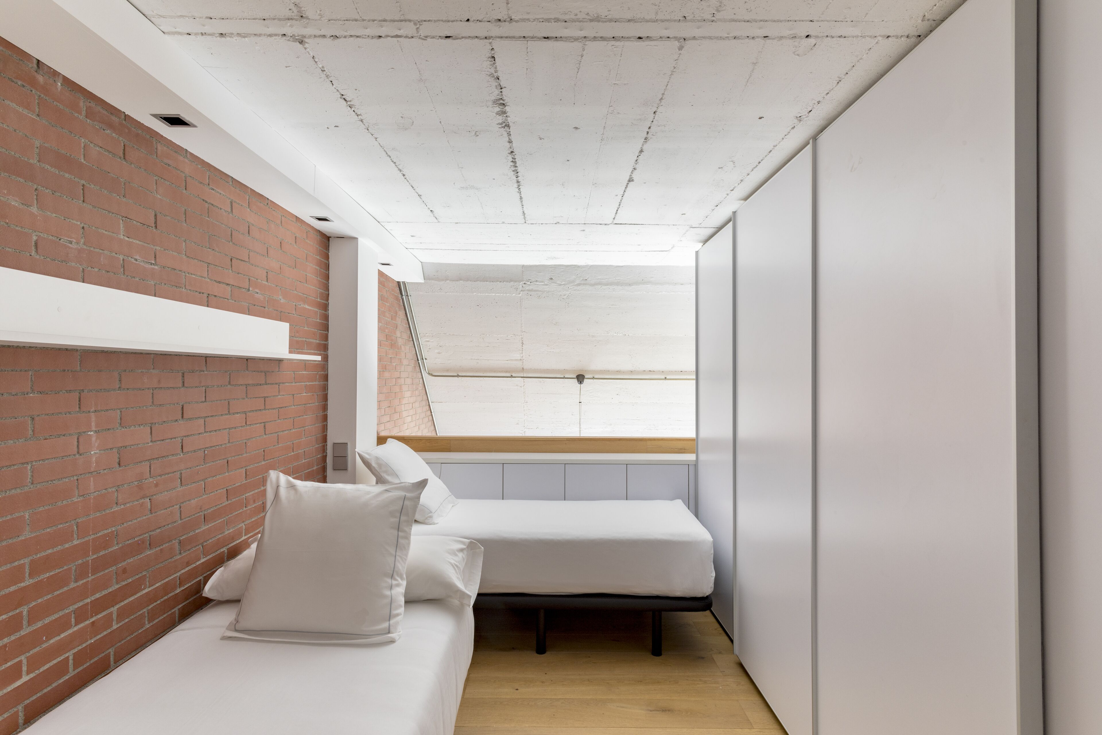 Une pièce de taille modeste avec un mur en briques, un lit blanc avec des oreillers, un plancher en bois et un plafond en pente doté d’une lucarne.